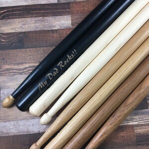 May include: A set of wooden drumsticks, including a pair of black drumsticks with the text "My Dad Rocks!" engraved on them.