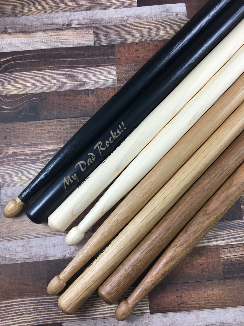 May include: A set of wooden drumsticks, including a pair of black drumsticks with the text "My Dad Rocks!" engraved on them.