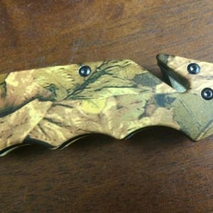Puede incluir: Cuchillo plegable con estampado de camuflaje, con una hoja y un cortador de cintur&oacute;n de seguridad. El mango presenta un dise&ntilde;o de hojas en tonos marr&oacute;n, verde y amarillo. El cuchillo tiene un acabado met&aacute;lico negro y est&aacute; dise&ntilde;ado para uso en exteriores.