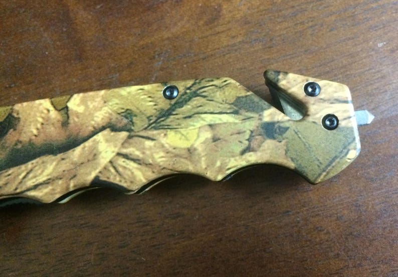 Puede incluir: Cuchillo plegable con estampado de camuflaje, con una hoja y un cortador de cintur&oacute;n de seguridad. El mango presenta un dise&ntilde;o de hojas en tonos marr&oacute;n, verde y amarillo. El cuchillo tiene un acabado met&aacute;lico negro y est&aacute; dise&ntilde;ado para uso en exteriores.