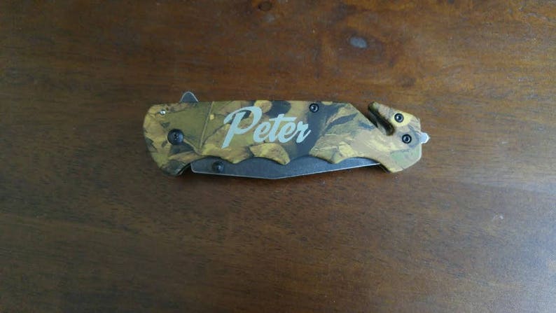 Puede incluir: Una navaja de bolsillo con estampado de camuflaje con el nombre "Peter" en cursiva. La navaja tiene una hoja negra y un cortador de seguridad incorporado. La navaja est&aacute; sobre una superficie de madera.