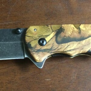 Puede incluir: Una navaja plegable con una hoja negra y un mango con estampado de camuflaje. La hoja es de metal oscuro y tiene un filo afilado. El mango presenta un dise&ntilde;o de camuflaje marr&oacute;n y beige con detalles en negro.