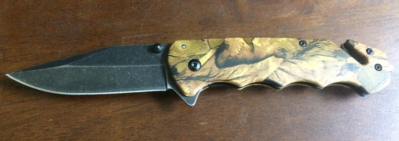 Puede incluir: Una navaja plegable con una hoja negra y un mango con estampado de camuflaje. La hoja es de metal oscuro y tiene un filo afilado. El mango presenta un dise&ntilde;o de camuflaje marr&oacute;n y beige con detalles en negro.