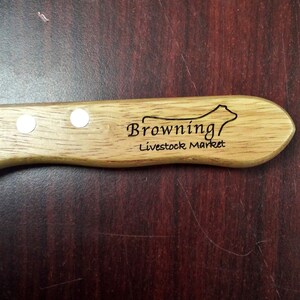 May include: A wooden-handled knife with a stainless steel blade. The handle is engraved with the text "Browning Livestock Market" and a silhouette of a cow.