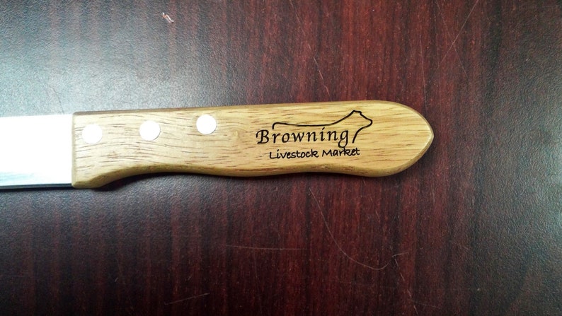 May include: A wooden-handled knife with a stainless steel blade. The handle is engraved with the text "Browning Livestock Market" and a silhouette of a cow.