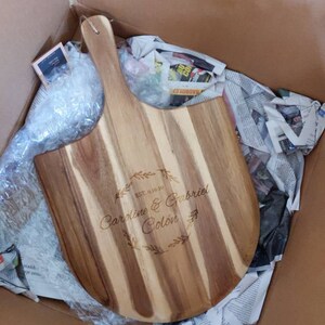 May include: A wooden pizza peel with a handle, personalized with the text "EST. 01.01.20 Caroline & Gabriel Colon" in a decorative font.