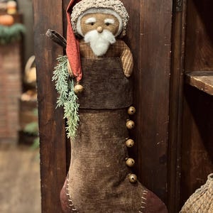 Peut inclure: Une chaussette de Noël en tissu marron ornée d'une figurine de Père Noël en peluche avec une barbe blanche et un bonnet rouge. La chaussette est décorée d'une rangée de petites clochettes en métal, d'un brin de pin vert et de coutures blanches.