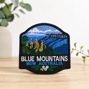 May include: A shield-shaped embroidered patch featuring a landscape of the Blue Mountains in Australia. The design includes golden rock formations, green trees, and blue mountains under a white sky. Text reads BLUE MOUNTAINS NSW AUSTRALIA.