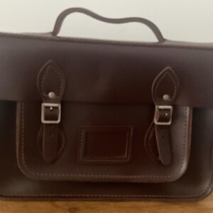 May include: A dark brown leather satchel bag with a top handle and a front flap secured by two silver buckles. The bag has a rectangular shape with a smaller front pocket and a stitched border. The bag is sitting on a wooden surface.