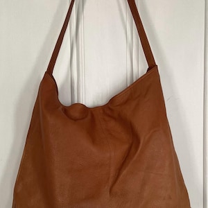 May include: A large, brown leather shoulder bag with long straps. The bag has a slouchy design and a smooth texture. The initials "C-S" are subtly embossed on the lower right corner. The bag is hanging against a white door.