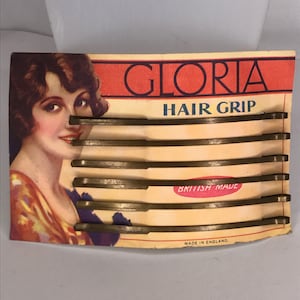 May include: A vintage card with a woman's illustration and the text "Gloria Hair Grip" in red letters. The card features six gold hair grips, labeled "British Made" and "Made in England".