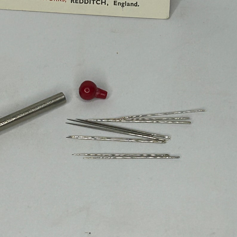 Puede incluir: Un juego de agujas de coser plateadas con un enhebrador de agujas de pl&aacute;stico rojo. Las agujas est&aacute;n dispuestas en una pila sobre una superficie blanca.