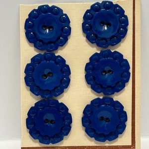 Peut inclure: Six boutons en plastique bleu avec un bord surélevé et deux trous. Les boutons sont disposés en deux rangées de trois sur une carte blanche.