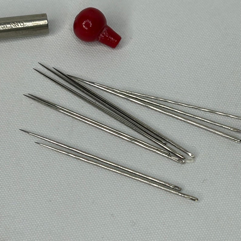 Puede incluir: Un juego de ocho agujas de coser plateadas con puntas afiladas y peque&ntilde;os agujeros para el hilo. Las agujas est&aacute;n dispuestas en forma de abanico sobre una superficie blanca.