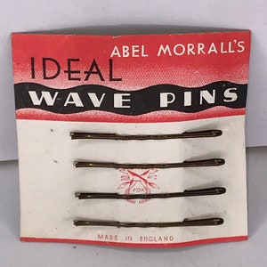 Op de afbeelding: Een kaart met vier gouden haarspeldjes om golven te maken. De kaart is rood en wit en zegt "ABEL MORRALL'S IDEAL WAVE PINS" in zwarte letters. De kaart zegt ook "MADE IN ENGLAND" in zwarte letters.