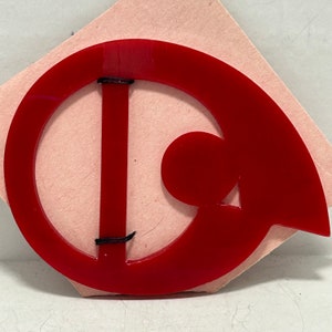 May include: A red plastic belt buckle with a unique design. The buckle is shaped like a letter D with a smaller circle cut out of the centre. The buckle has two black straps attached to it.