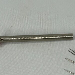Puede incluir: Un estuche para agujas de metal plateado con el texto "SHIMETONS NEEDLE CASE MADE IN ENGLAND" grabado en el lateral.