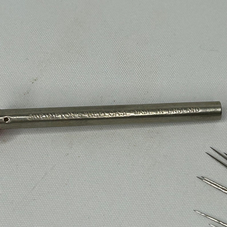 Puede incluir: Un estuche para agujas de metal plateado con el texto "SHIMETONS NEEDLE CASE MADE IN ENGLAND" grabado en el lateral.