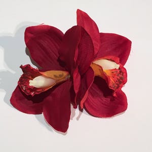 May include: Two red silk orchid flowers with yellow and orange centres. The flowers are arranged together and have a slightly ruffled texture.