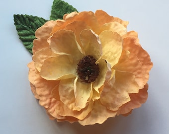 May include: A large, artificial flower with layered peach and yellow petals and a dark brown center. Two green felt leaves are attached to the flower. The flower is a decorative item.