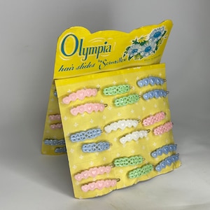 May include: A display card with the text "Olympia by Scintilles hair slides" and a variety of colourful plastic hair slides with heart designs. Made in England.