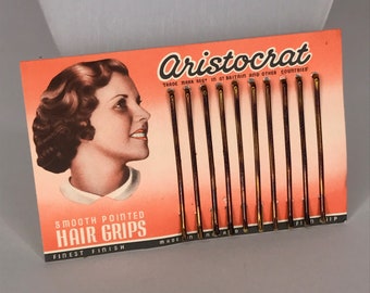 Könnte beinhalten: Eine Vintage-Karte mit Haarklammern namens "Aristocrat" mit dem Gesicht einer Frau auf der Vorderseite. Die Karte ist rot und weiß mit goldenen Buchstaben. Die Haarklammern sind glatt und spitz und wurden in England hergestellt. Der Text auf der Karte lautet "Smooth Pointed Hair Grips Finest Finish".
