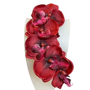 May include: A cascade of deep red orchid flowers with dark pink accents, artfully arranged on a white, curved display. The petals have a velvety texture, creating a striking contrast against the smooth, neutral backdrop. This floral arrangement is ideal for home decor.