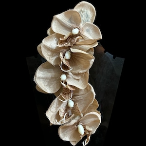 May include: A vertical arrangement of beige orchid flowers with white and brown accents. The flowers are displayed on a black stand against a black background. The orchid arrangement is a decorative floral piece.