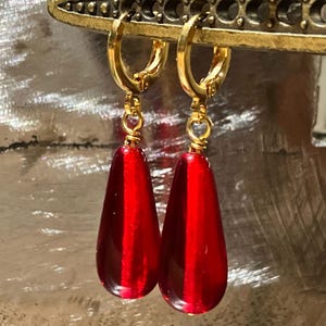 May include: A pair of gold-colored hoop earrings with teardrop-shaped, translucent red pendants. The earrings have small heart-shaped accents and are displayed against a textured, silver background. The earrings are a jewelry item.