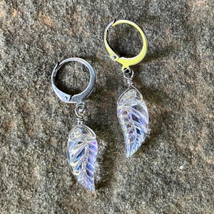 May include: A pair of silver-toned hoop earrings featuring dangling, iridescent, clear glass leaf-shaped charms. Each leaf charm is attached to the hoop with a small silver wire wrap, displaying a textured vein pattern and a subtle rainbow sheen.