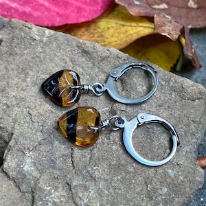May include: A pair of earrings featuring heart-shaped amber-colored glass beads with black stripes. Each bead is attached to a silver-toned metal leverback hoop by a small wire-wrapped loop, creating a dangling design for these earrings.