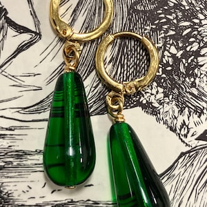 May include: A pair of gold-toned hoop earrings featuring dangling, teardrop-shaped green glass beads. Each bead has a smooth, polished finish with subtle, darker horizontal line patterns visible within the translucent green glass structure.