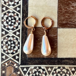 May include: A pair of gold-colored hoop earrings with pearlescent, teardrop-shaped pendants. The earrings are displayed on a decorative wooden surface with geometric patterns in shades of brown, beige, and black.