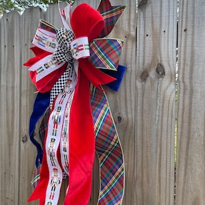 May include: A festive Christmas bow with a variety of ribbons in red, green, blue, and white. The bow features a large red velvet center, plaid accents, and ribbons with nutcracker soldier designs. It is attached to a wooden fence.