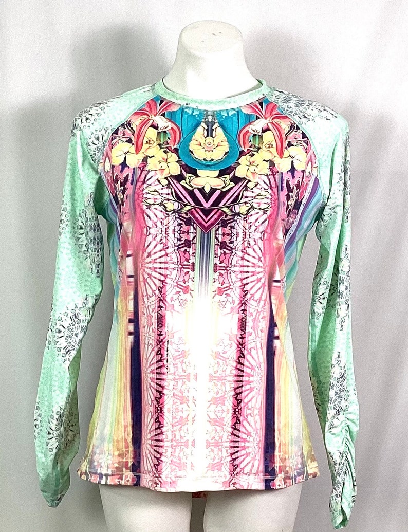 Op de afbeelding: Een shirt met lange mouwen met een kleurrijke, abstracte print met bloemen en geometrische patronen. Het shirt heeft een lichtgroene basiskleur met een wit en zilveren patroon op de mouwen.