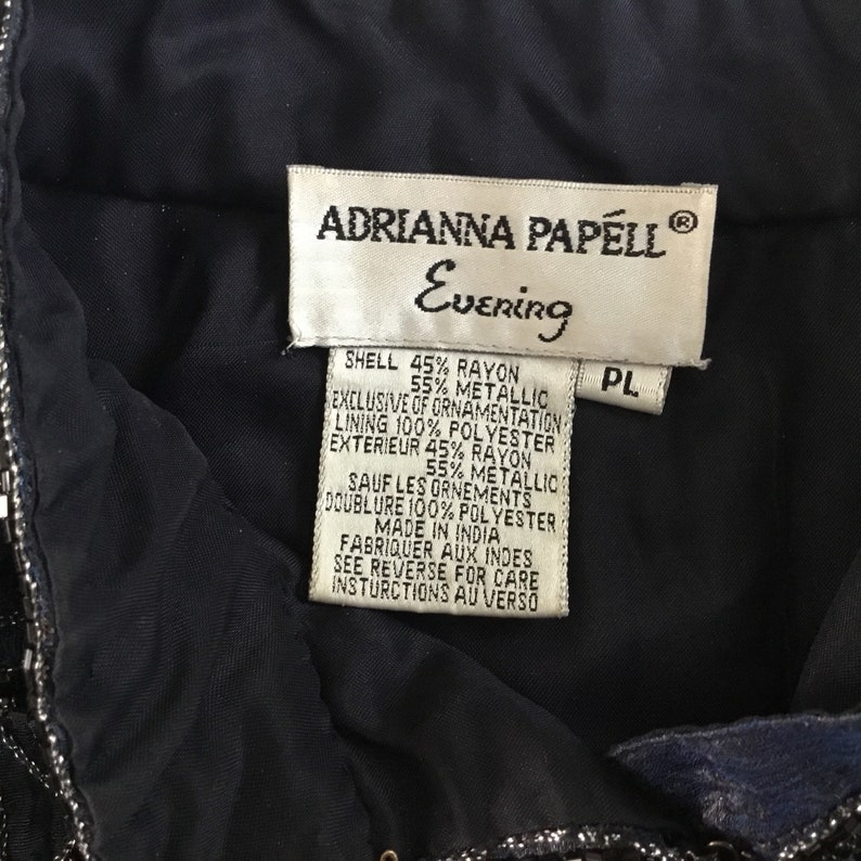 K&ouml;nnte beinhalten: Nahaufnahme eines Etiketts von einem Adrianna Papell Evening Kleidungsst&uuml;ck. Das Etikett ist wei&szlig; mit schwarzem Text, der die Materialien von Oberstoff und Futter auflistet. Das Kleidungsst&uuml;ck ist dunkelblau mit silbernen Verzierungen.