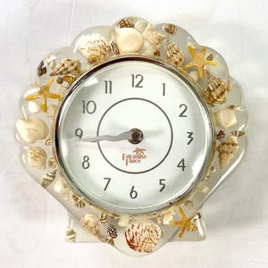 May include: A seashell-shaped clock with a white face and silver trim. The clock is embedded with various seashells and starfish. The clock face has black numerals and the words "Paradise Place".