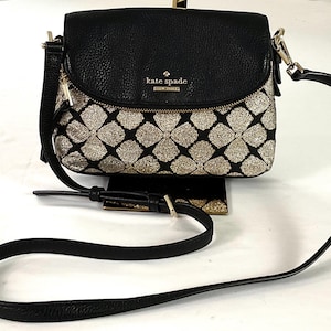 May include: Black leather crossbody bag with a flap closure. The lower portion of the bag features a gold glitter pattern. The bag has a long, adjustable strap and a gold-toned zipper and hardware. The brand name "kate spade" is visible.