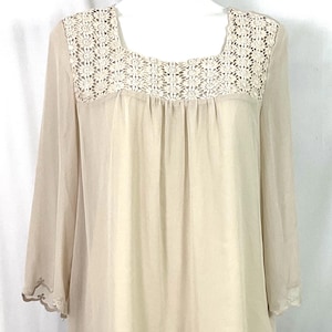 May include: A beige blouse with a square neckline and long sleeves. The neckline features a detailed, cream-colored lace design. The blouse has a flowing, semi-sheer fabric and scalloped edges on the sleeves and hemline.