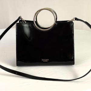 May include: A black patent leather handbag with a rectangular shape, featuring two circular silver-tone metal top handles and a detachable shoulder strap. The front displays the gold-tone text Kate Spade New York centered near the bottom edge.