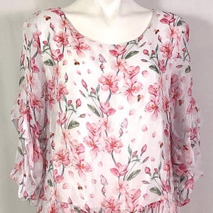 May include: A white blouse featuring a delicate pink cherry blossom print with green leaves and small bees. The garment has a round neckline, three-quarter length sleeves with ruffled edges, and a ruffled hemline, creating a soft, layered appearance.