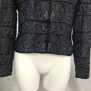 K&ouml;nnte beinhalten: Marineblaue Jacke mit aufwendigen schwarzen, perlenbesetzten Blumen- und geometrischen Mustern. Die Jacke hat lange &Auml;rmel und eine Knopfleiste. Das Design umfasst horizontale B&auml;nder und detaillierte Verzierungen, die einen strukturierten und eleganten Look erzeugen.