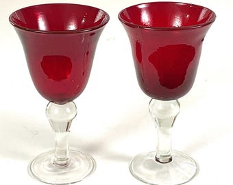 May include: Two ruby red wine glasses. The bowls are a deep red, transitioning to clear stems and bases. The glasses have a classic tulip shape, perfect for serving wine. The clear stems and bases provide a striking contrast to the red bowls.