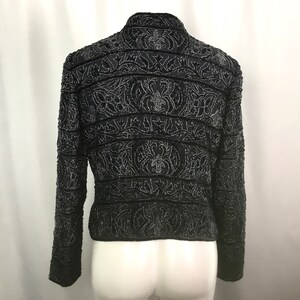 K&ouml;nnte beinhalten: Schwarze, mit Perlen besetzte Jacke mit aufwendigen floralen und geometrischen Mustern. Die Jacke hat lange &Auml;rmel und einen Stehkragen. Das Design umfasst horizontale B&auml;nder und detaillierte Verzierungen, die einen strukturierten und eleganten Look erzeugen. Die Jacke wird auf einer Schaufeniquin pr&auml;sentiert.