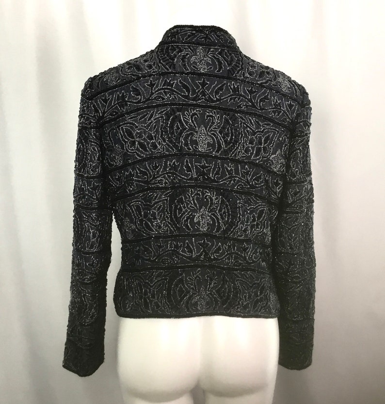 K&ouml;nnte beinhalten: Schwarze, mit Perlen besetzte Jacke mit aufwendigen floralen und geometrischen Mustern. Die Jacke hat lange &Auml;rmel und einen Stehkragen. Das Design umfasst horizontale B&auml;nder und detaillierte Verzierungen, die einen strukturierten und eleganten Look erzeugen. Die Jacke wird auf einer Schaufeniquin pr&auml;sentiert.