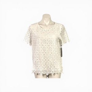 May include: A white, short-sleeved top with a delicate, openwork crochet design. The top features a round neckline and scalloped edges on the sleeves and hem. The intricate pattern creates a textured, airy look. A tag hangs from the side.