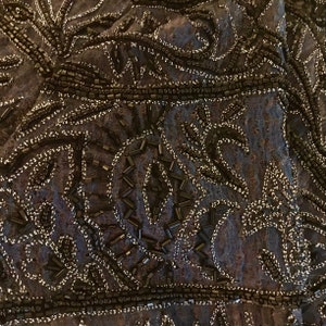 K&ouml;nnte beinhalten: Nahaufnahme eines dunkelblauen Stoffes, der reich mit schwarzen Perlen und silbernem Faden verziert ist. Das Design zeigt komplizierte Blumen- und geometrische Muster, die ein strukturiertes und verziertes Aussehen erzeugen. Die Perlen sind dicht angeordnet.
