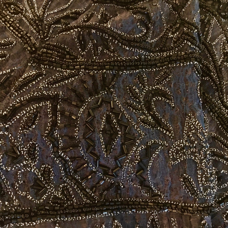 K&ouml;nnte beinhalten: Nahaufnahme eines dunkelblauen Stoffes, der reich mit schwarzen Perlen und silbernem Faden verziert ist. Das Design zeigt komplizierte Blumen- und geometrische Muster, die ein strukturiertes und verziertes Aussehen erzeugen. Die Perlen sind dicht angeordnet.