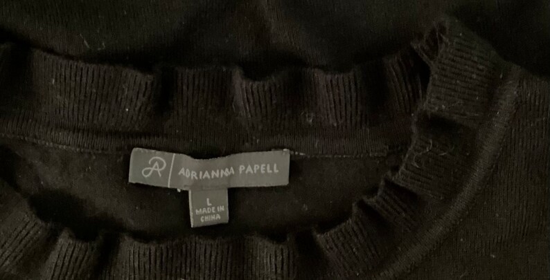 K&ouml;nnte beinhalten: Schwarzer Pullover mit R&uuml;schenkragen und einem Etikett mit der Aufschrift "Adrianna Papell" und "L" f&uuml;r die Gr&ouml;&szlig;e. Das Etikett tr&auml;gt au&szlig;erdem die Aufschrift "Made in China".
