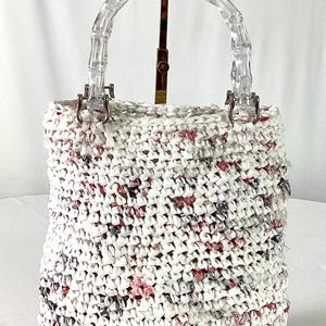 May include: A white crocheted tote bag with a clear, bamboo-style handle. The bag is made of white material with flecks of red, pink, and blue. The bag is sitting on a gold-colored stand.
